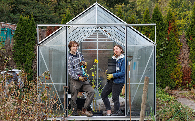 Two students pose in a glasshouse in a community garden.One holds a shovel. The other holds pots.